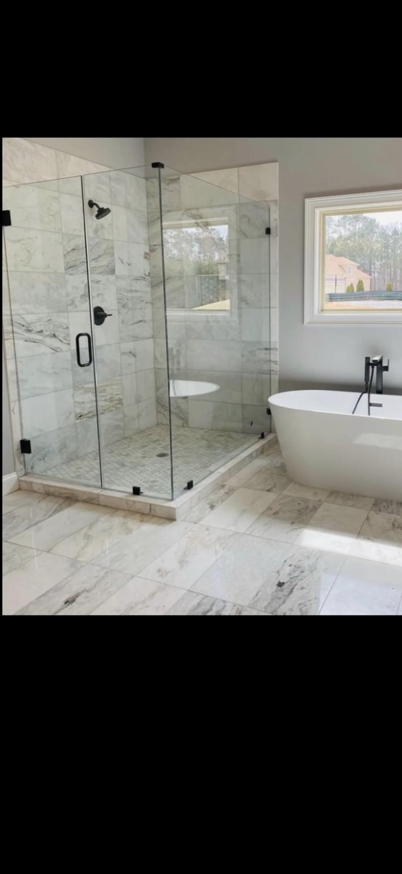 Frameless glass shower enclosure with black hardware in marble bathroom with freestanding tub